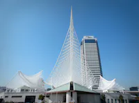 A modern white steel architectural structure with a tall, pointed spire stands in front of a high-rise building, set against a clear blue sky.