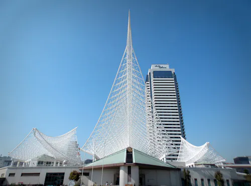 A modern white steel architectural structure with a tall, pointed spire stands in front of a high-rise building, set against a clear blue sky.