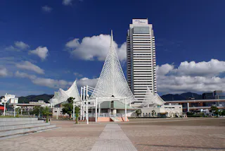 A modern cityscape features a tall, white skyscraper and unique, sail-like architectural structures under a blue sky with scattered clouds, with a wide open plaza in the foreground.
