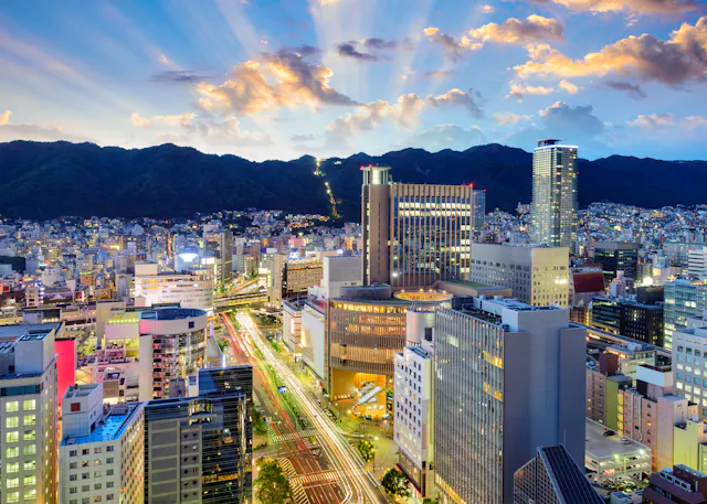 Cityscape at sunset with tall buildings and busy streets in the foreground, mountains in the background, and sun rays shining through colorful clouds in the sky.
