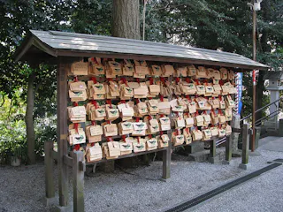 A wooden board at a Japanese shrine displays many ema plaques, each with handwritten prayers or wishes, hanging by red strings. The area is shaded by trees, and gravel covers the ground.
