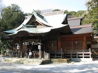 A traditional Japanese Shinto shrine with ornate wooden architecture, a curved roof, and decorative lanterns at the entrance, surrounded by trees and a gravel courtyard. Several people stand at the entrance steps.