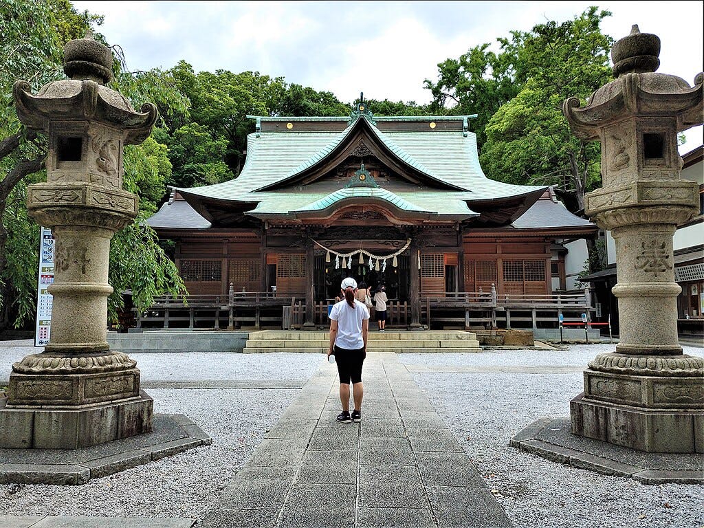 A person stands facing a traditional Japanese shrine, flanked by two large stone lanterns, with greenery in the background and a gravel courtyard in the foreground.