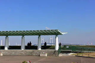 A pergola with white columns overlooks a distant suspension bridge and cranes under a clear blue sky. Several people stand or walk near the structure on a paved area with planters and railings.