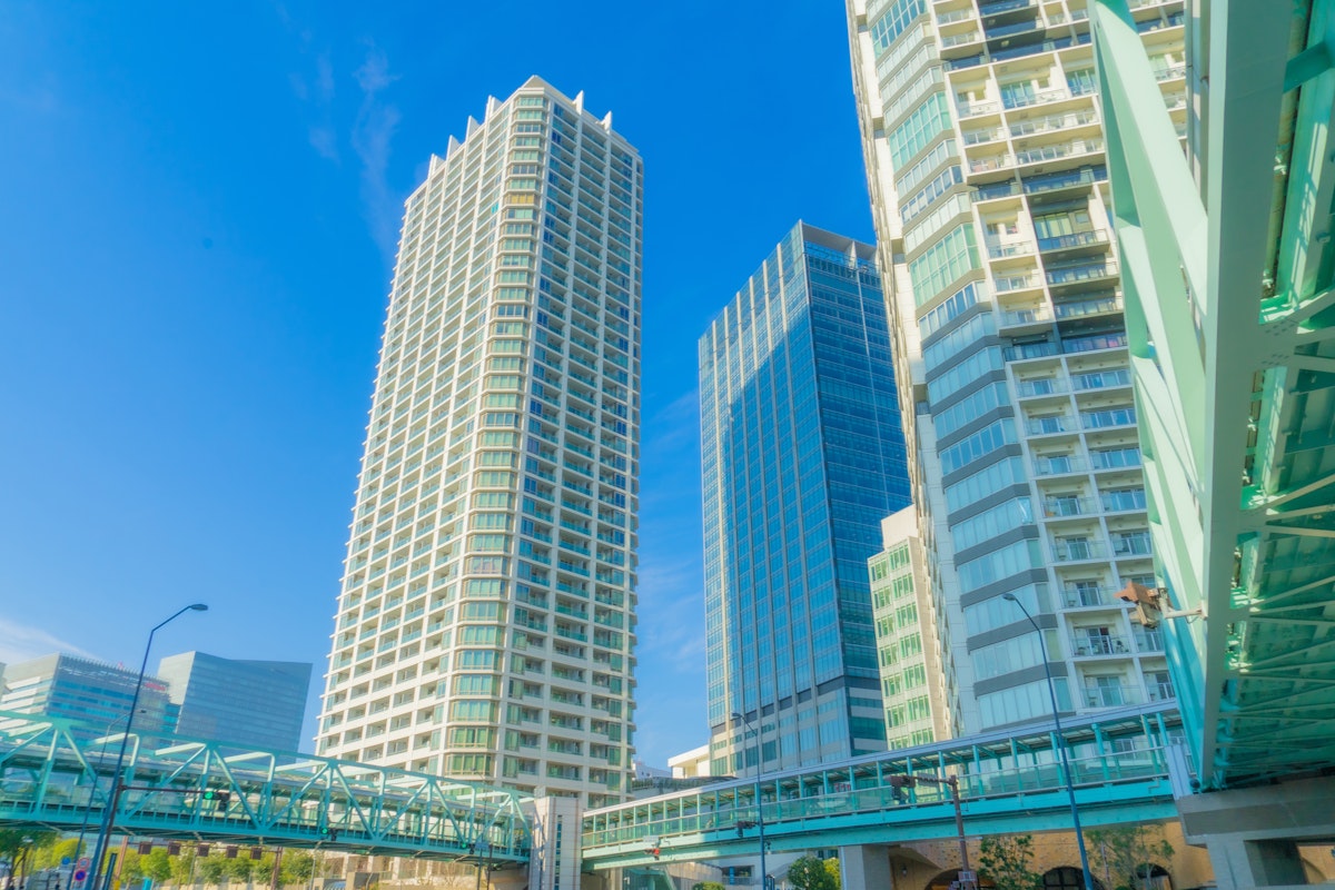 Yokohama Weather A vibrant cityscape featuring several tall, modern high-rise buildings against a clear blue sky. Green steel pedestrian bridges connect the structures at varying heights, while more buildings are visible in the background. Sunlight brightens the scene.