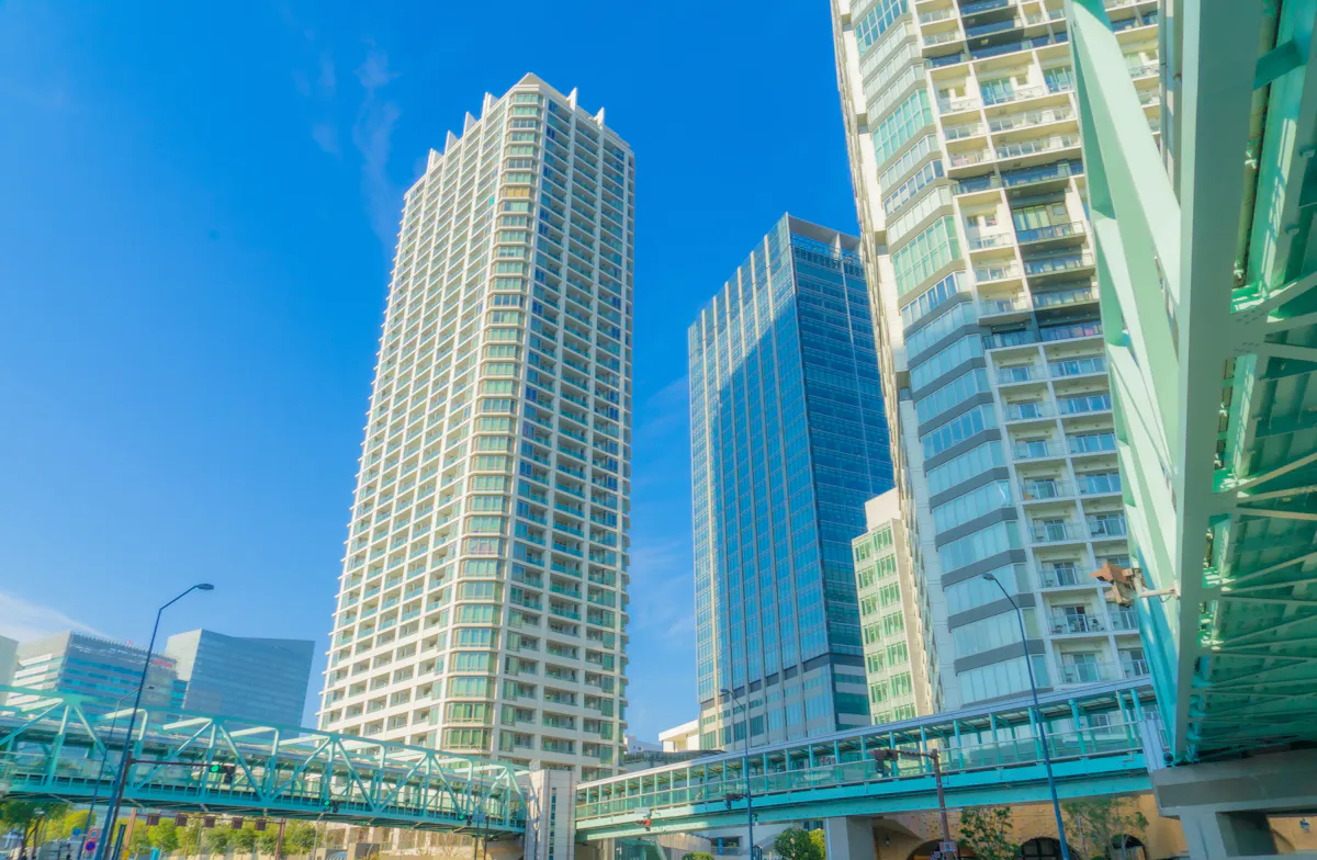 A vibrant cityscape featuring several tall, modern high-rise buildings against a clear blue sky. Green steel pedestrian bridges connect the structures at varying heights, while more buildings are visible in the background. Sunlight brightens the scene.
