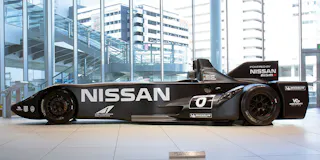 A sleek black Nissan race car with "NISSAN" in large white letters on its side is displayed indoors, surrounded by glass windows and modern buildings outside.
