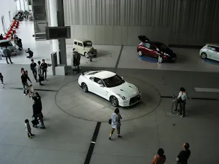 A white sports car is displayed on a circular platform indoors, surrounded by people. Other classic and modern cars are visible in the background of the spacious, well-lit exhibition hall.