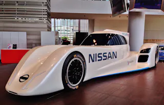 A sleek, futuristic white Nissan race car with aerodynamic design and black accents is displayed indoors on a wooden floor, featuring prominent "NISSAN" branding on its side.