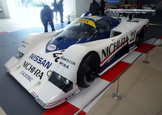 A Nissan race car with a white, blue, and red livery, number 23, and prominent “NICHIRA” and “NISSAN” logos, displayed indoors behind a barrier, with people viewing in the background.