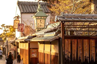 A sunlit street in Japan features traditional wooden buildings, tiled roofs, hanging signs, and a green lantern. A few people walk along the narrow path, with trees and warm golden light enhancing the serene atmosphere.