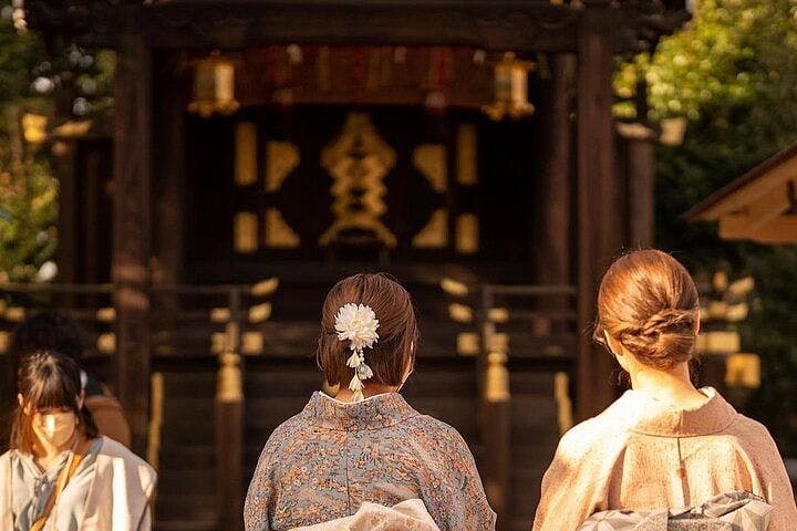 Three women in traditional Japanese clothing stand with their backs to the camera, facing a wooden shrine in soft sunlight. One woman has a white flower hair accessory. Trees and golden light frame the scene.
