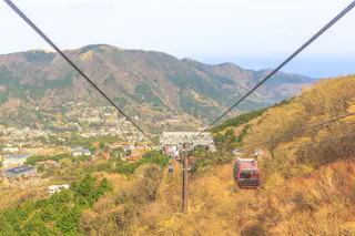 Red cable cars travel up and down a mountain, suspended by cables above a lush, green valley with a town below and forested hills in the background under a partly cloudy sky.