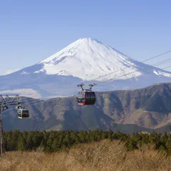 Hakone Ropeway Two cable cars travel above a forested area with Mount Fuji, partially covered in snow, towering in the background under a clear blue sky.