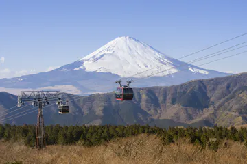 Hakone Ropeway Two cable cars travel above a forested area with Mount Fuji, partially covered in snow, towering in the background under a clear blue sky.