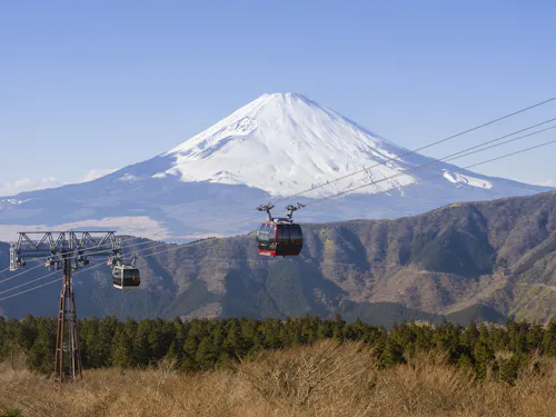 Hakone Ropeway Two cable cars travel above a forested area with Mount Fuji, partially covered in snow, towering in the background under a clear blue sky.