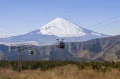 Two cable cars travel above a forested area with Mount Fuji, partially covered in snow, towering in the background under a clear blue sky.