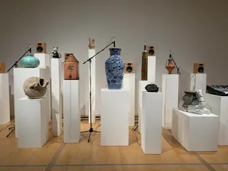 A variety of ceramic vessels and sculptures are displayed on white pedestals, surrounded by microphones and speakers in a gallery setting with a wooden floor and neutral walls.
