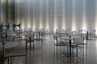 A modern, minimalist indoor café with round tables and metallic chairs, each table adorned with a single red flower. The backdrop features a textured, lighted wall, creating a sleek, contemporary atmosphere.