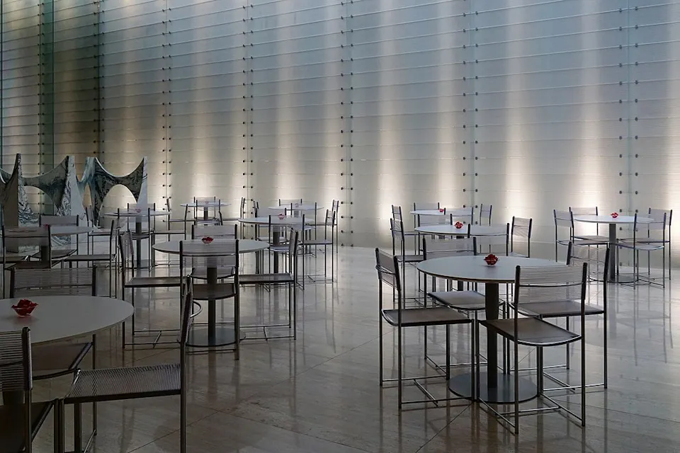 A modern, minimalist indoor café with round tables and metallic chairs, each table adorned with a single red flower. The backdrop features a textured, lighted wall, creating a sleek, contemporary atmosphere.