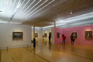 People view framed paintings displayed on white and pink walls in a spacious, well-lit art gallery with wooden floors and a modern ceiling.