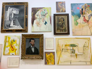 A gallery wall displays various abstract and cubist-style paintings and sketches, including portraits, figures, and a cat, all in assorted frames and sizes against a white background.