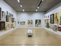 A brightly lit art gallery features white walls displaying numerous colorful paintings, mostly portraits and abstract art. A white bench sits in the center of the light wood floor.