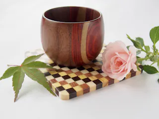 A polished wooden cup sits on a checkered cutting board next to a pink rose and a green maple leaf, all arranged on a white background.