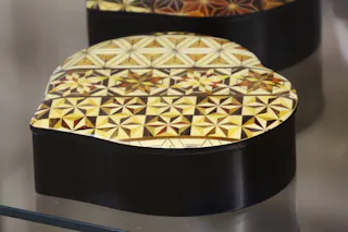 A heart-shaped box with a black base and a lid decorated with intricate geometric patterns in shades of yellow, brown, and cream, displayed on a reflective surface.