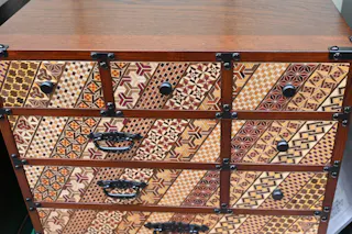 A wooden chest of drawers featuring intricate geometric patterns in various colors and designs on each drawer, with black metal handles and corner accents.