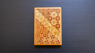 A rectangular wooden box with intricate geometric patterns in shades of brown, tan, and beige, displayed on a dark, ridged surface.