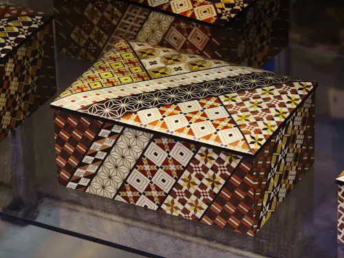 Hakone-Yosegi-Zaiku A decorative wooden box with a hinged lid, covered in intricate geometric mosaic patterns in shades of brown, yellow, white, and black. The patterns feature diamonds, squares, and star designs.