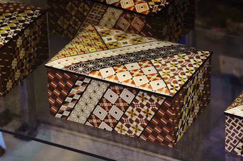 A decorative wooden box with a hinged lid, covered in intricate geometric mosaic patterns in shades of brown, yellow, white, and black. The patterns feature diamonds, squares, and star designs.