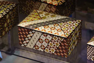 A decorative wooden box with a hinged lid, covered in intricate geometric mosaic patterns in shades of brown, yellow, white, and black. The patterns feature diamonds, squares, and star designs.