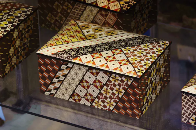 A decorative wooden box with a hinged lid, covered in intricate geometric mosaic patterns in shades of brown, yellow, white, and black. The patterns feature diamonds, squares, and star designs.