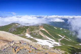 Hikers walk along a rocky mountain trail with patches of snow, surrounded by green hills and a winding road. Clouds hang low in the blue sky, partially covering distant mountain peaks.