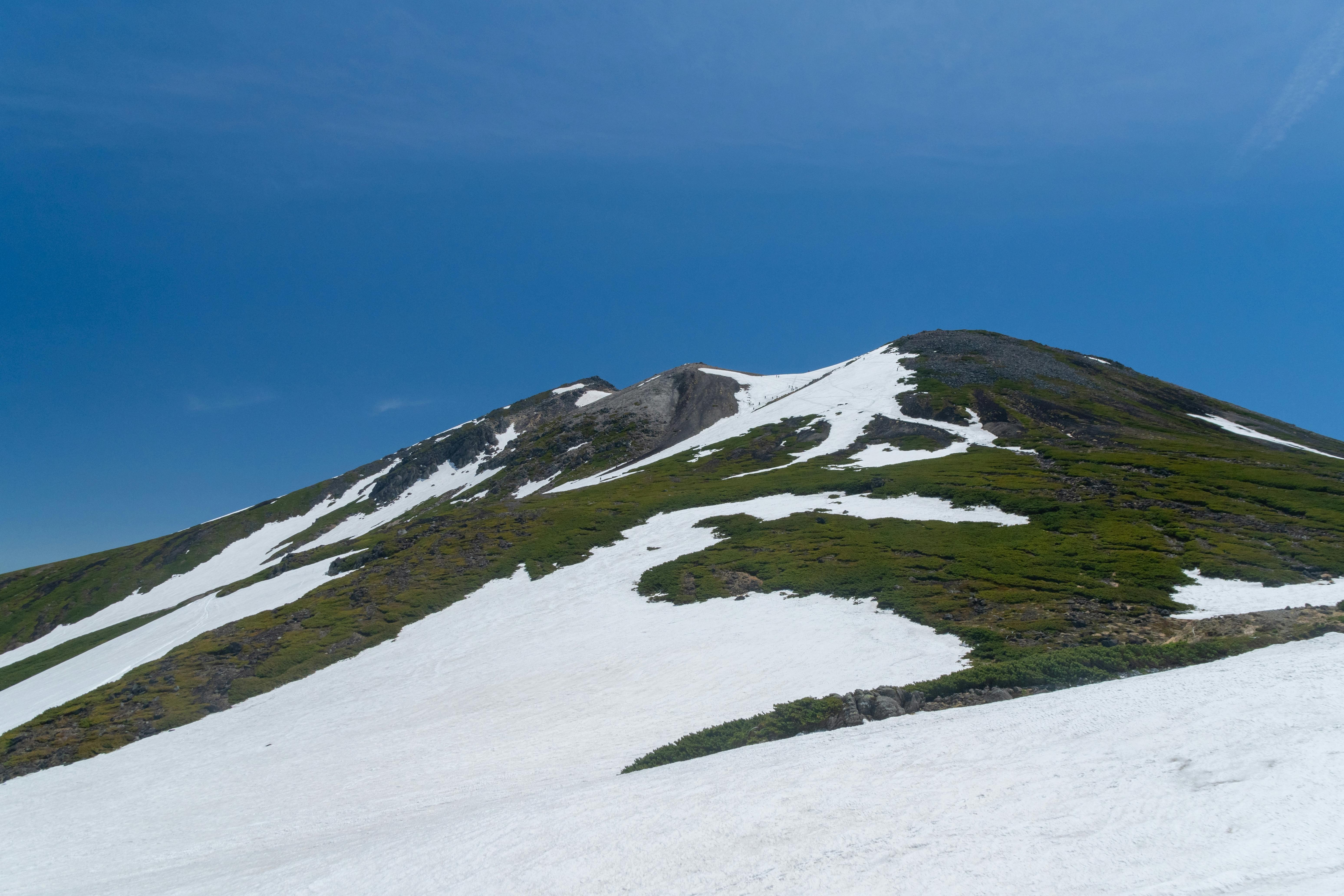 A mountain slope partially covered with patches of snow and green vegetation under a clear blue sky.