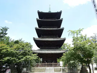 A traditional five-story Japanese pagoda stands surrounded by trees, with its tiered roofs and intricate wooden architecture visible under a bright, partly cloudy sky.