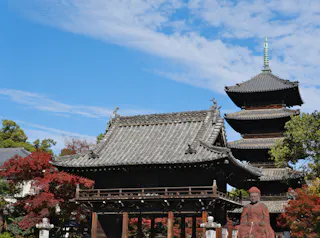 A traditional Japanese temple with a tiled roof, a tall pagoda in the background, autumn trees with red leaves, and a red statue in the foreground under a blue sky with wispy clouds.