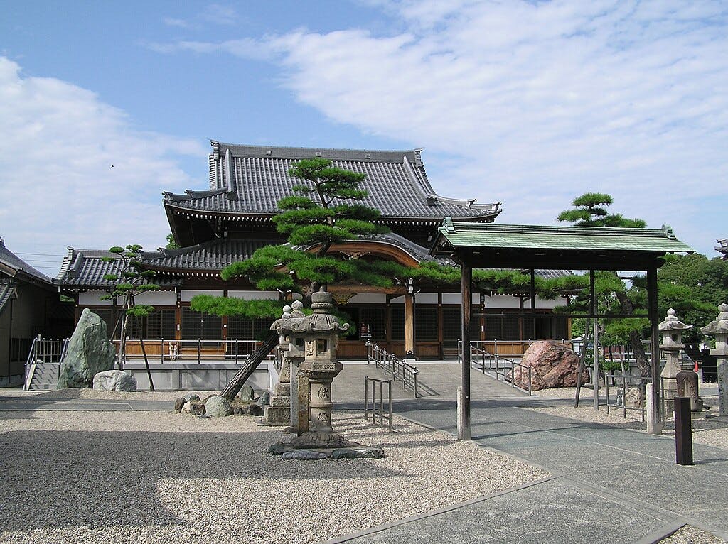A traditional Japanese temple with a tiled roof, surrounded by neatly trimmed trees, stone lanterns, and a gravel courtyard under a partly cloudy sky.
