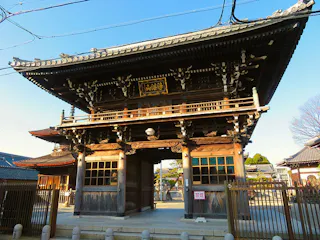 A traditional Japanese wooden temple gate with intricate carvings and a tiled roof, surrounded by a fence and illuminated by sunlight, with clear blue sky in the background.