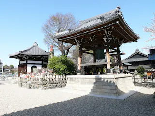 A traditional Japanese temple courtyard with a wooden bell tower, stone statues, and a tiled-roof building, surrounded by trees and gravel under a clear blue sky.