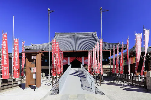 Traditional Japanese temple with a sloped roof, surrounded by tall red banners with white text. A ramp leads to the entrance, and the sky above is bright blue and clear.