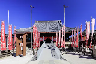 Traditional Japanese temple with a sloped roof, surrounded by tall red banners with white text. A ramp leads to the entrance, and the sky above is bright blue and clear.