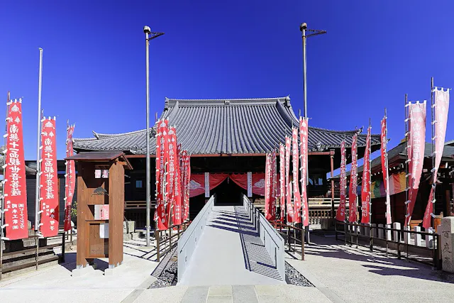 Traditional Japanese temple with a sloped roof, surrounded by tall red banners with white text. A ramp leads to the entrance, and the sky above is bright blue and clear.