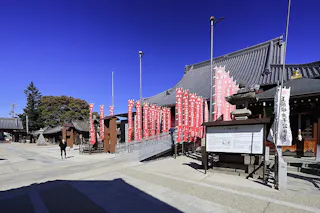 A traditional Japanese temple with tiled roofs, red banners with white writing, stone lanterns, and informational signs, set against a clear blue sky. A few people walk near the buildings.