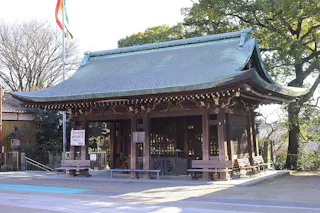 A traditional Japanese Shinto shrine with a curved roof and wooden structure, surrounded by trees and flags, bathed in sunlight. Benches are placed in front of the shrine, and shadows fall across the paved ground.