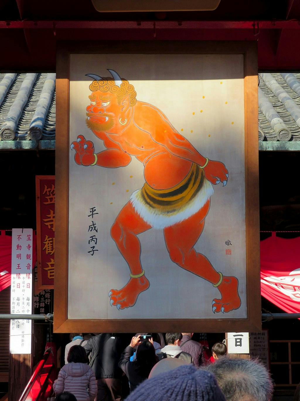 A colorful painting of a red-skinned, horned oni (demon) with sharp claws and fangs, wearing a straw loincloth, is displayed above a crowd of people at a shrine or temple.