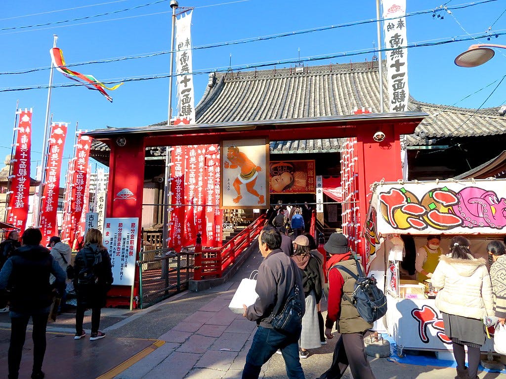 People walk near a vibrant red Japanese temple gate adorned with banners and flags. Food stalls and colorful signs are visible, suggesting a festive or market atmosphere on a sunny day.