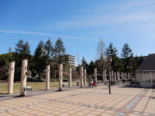 A spacious outdoor plaza with tall, cylindrical columns, benches, potted flowers, and people sitting. Trees and a few buildings are in the background under a clear blue sky.